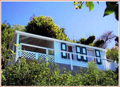 A charming white structure is nestled amidst lush greenery, featuring multiple green-shuttered windows and a balcony with white railings. The bright blue sky provides a vivid backdrop, complementing the tropical atmosphere.