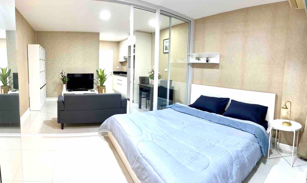 A spacious bedroom features a large bed with a blue comforter and decorative pillows. A modern sofa is placed adjacent, along with a side table and a lamp. The room is divided by glass panels, creating an open feel and allowing light to illuminate the space.