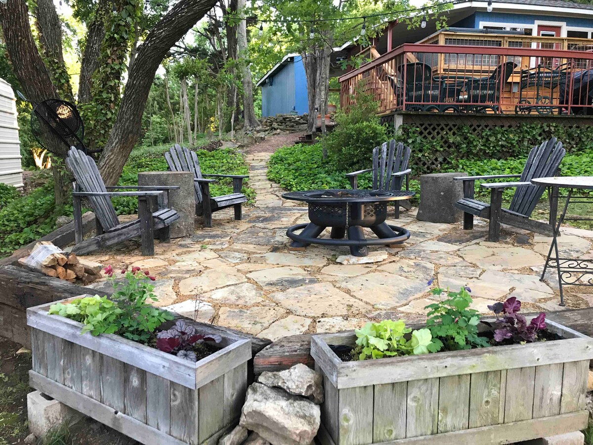An outdoor seating area is surrounded by stone paving, featuring several wooden Adirondack chairs arranged around a central fire pit. Planters with various green plants are placed along the edge, contributing to the natural ambiance of the area nestled among trees.