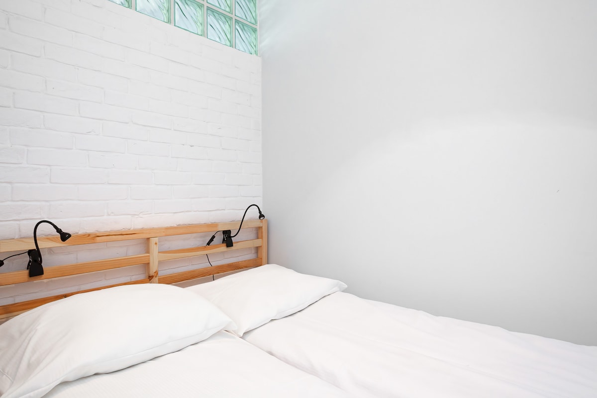 简约的卧室，配有白色砖墙、舒适的双人床和明亮的自然光线，营造出宁静祥和的氛围。