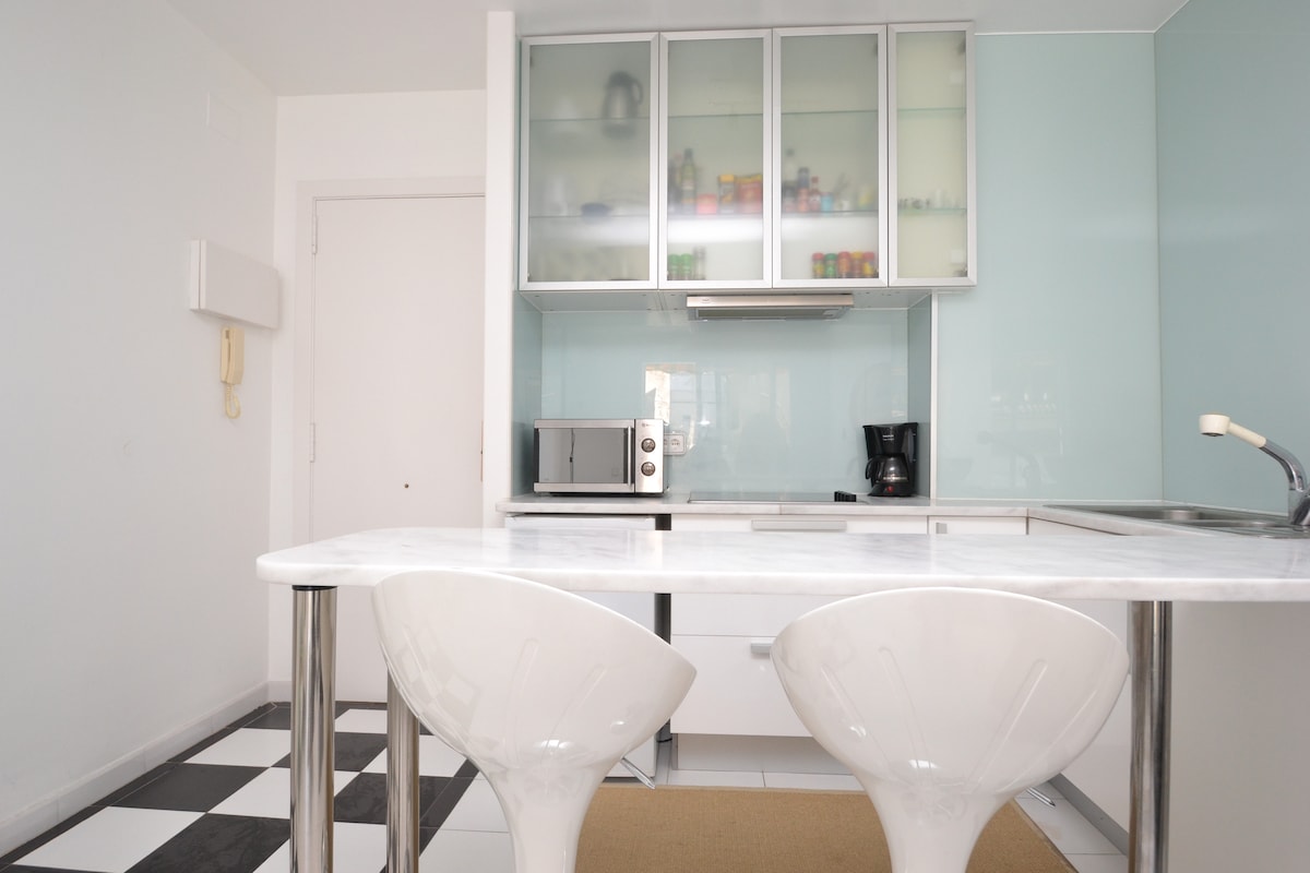 The kitchen area features sleek cabinetry with a light blue backdrop. A marble countertop is accompanied by two white, modern bar stools. Appliances, including a microwave and coffee maker, are visible, with additional shelves displaying various items in organized fashion.