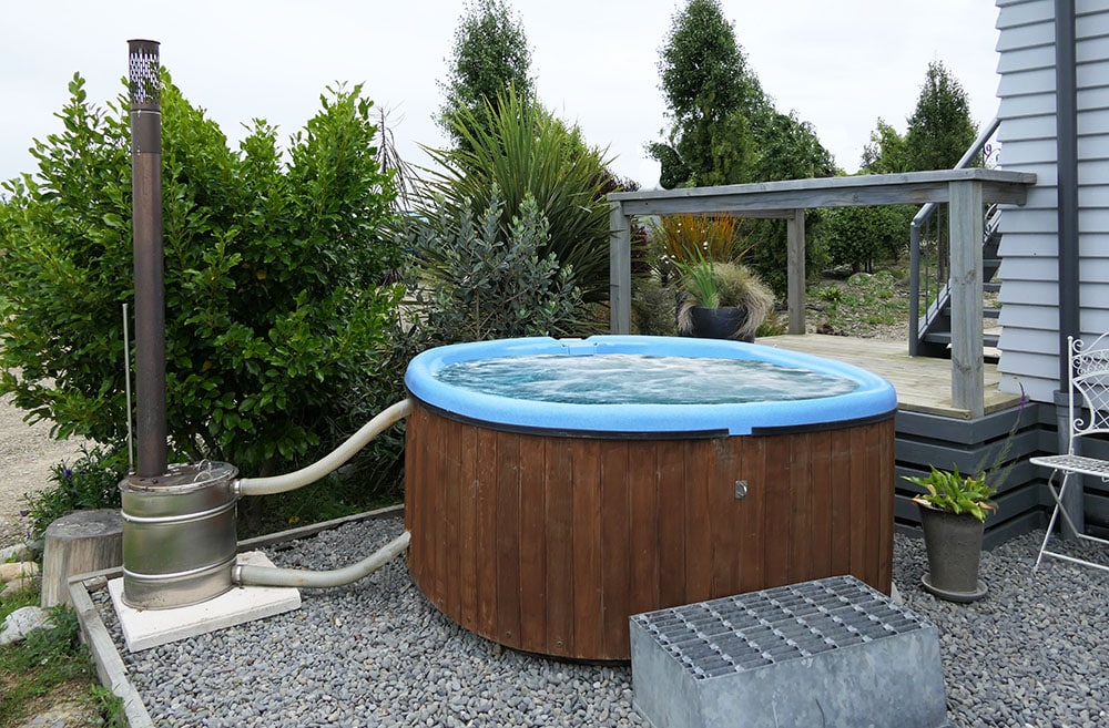 A wooden spa pool sits outdoors, surrounded by greenery and gravel. A metal stove is positioned nearby, with a small bench and potted plant adding to the outdoor space. The spa is filled with water, displaying inviting blue edges.