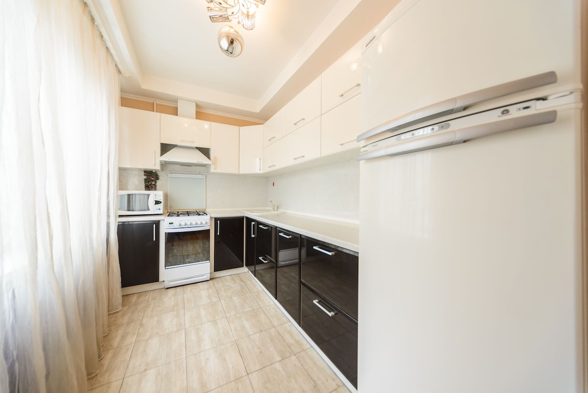 The kitchen features modern cabinetry with a mix of dark and light colors, providing a sleek appearance. Equipped with an oven, stove, microwave, and refrigerator, the space offers essential appliances. Large windows allow natural light to fill the area, enhancing the inviting ambiance.