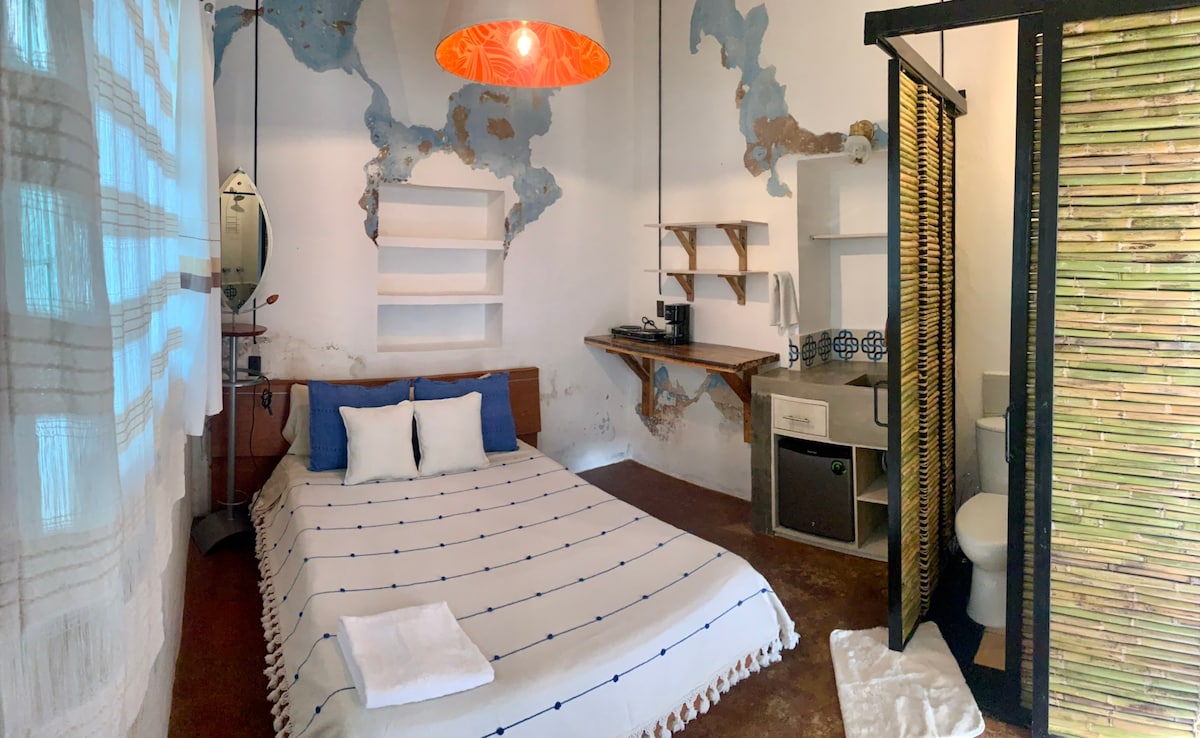 A spacious sleeping area features a large bed covered with a white blanket adorned with blue accents. Exposed walls reveal a textured finish, while open shelving and a counter area are visible. A partitioned section displays a modern bathroom with essential fixtures.