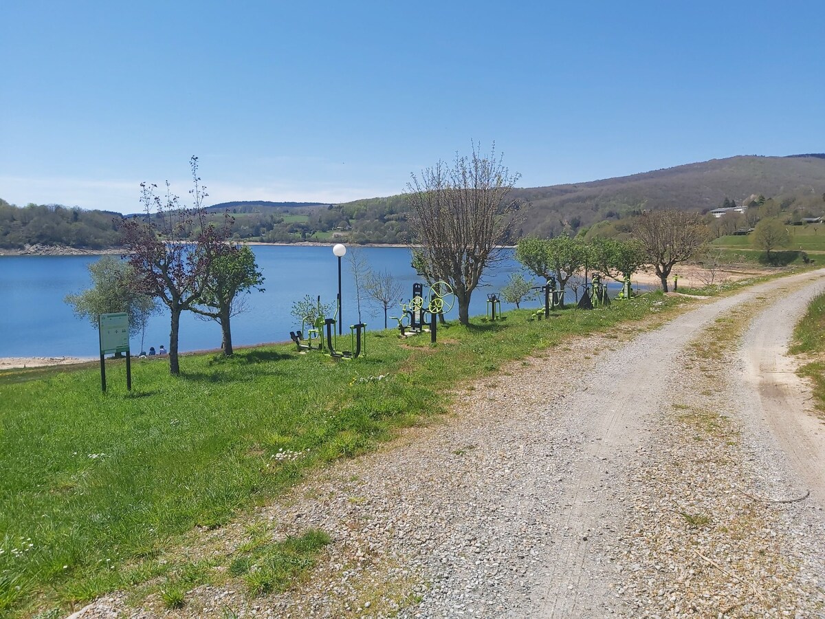 A scenic pathway lined with grass and trees leads to the sparkling water of Lake Laouzas. Recreational equipment is situated along the shore, providing opportunities for outdoor activities. The surrounding landscape features gentle hills under a clear blue sky, creating a peaceful atmosphere.