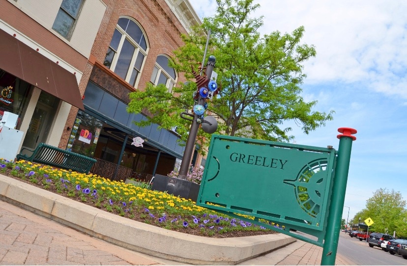 A pedestrian-friendly street in Greeley features a decorative sign alongside vibrant flower beds filled with blooms. Nearby, a tree provides greenery against a backdrop of historic brick architecture. Outdoor seating is visible, contributing to a welcoming urban atmosphere.