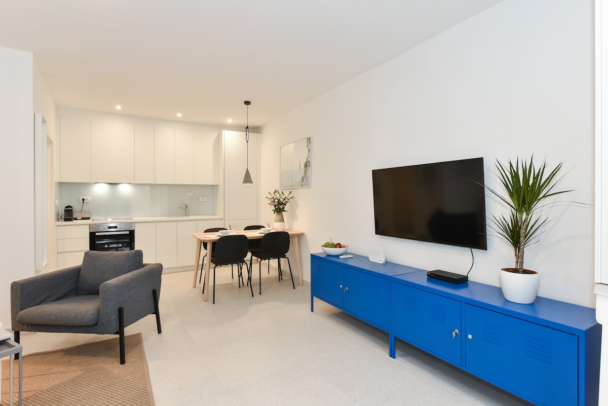 A modern open living space features a sleek blue media unit below a wall-mounted television. A small dining area with four black chairs is visible next to a contemporary kitchen with white cabinetry. A potted plant adds a touch of greenery to the room.