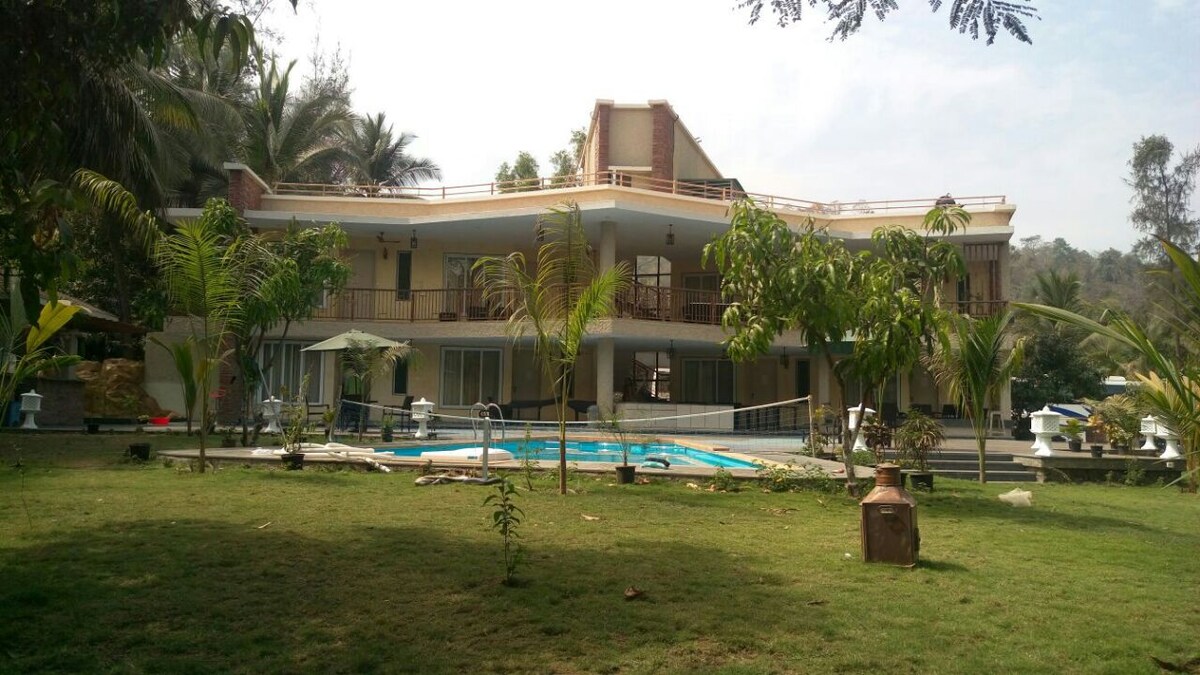 A spacious estate is presented, featuring multiple levels with large glass windows. The swimming pool is centrally located, surrounded by lush greenery and palm trees. Comfortable seating areas are visible, providing a relaxed outdoor environment for guests to enjoy.