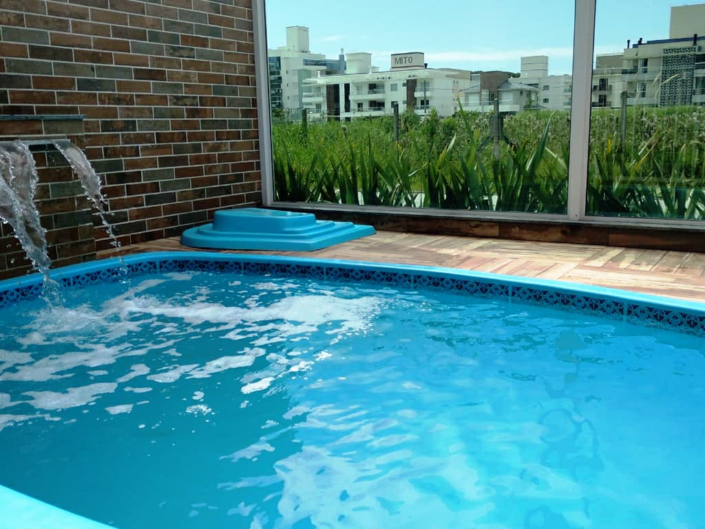 A bright and inviting shared pool area is highlighted by clear blue water, with a gentle waterfall feature on one side. The surrounding glass walls offer views of lush greenery and city buildings in the distance, creating a serene atmosphere.