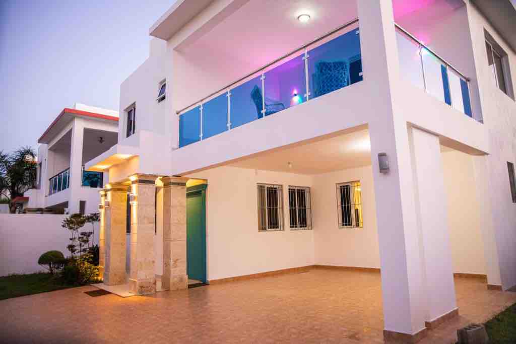 A modern two-story house is shown from the exterior, featuring a spacious patio area with tiled flooring. The entrance is highlighted by recessed lighting, and large windows provide glimpses into the interior. A balcony is visible on the upper level, adorned with glass railings.