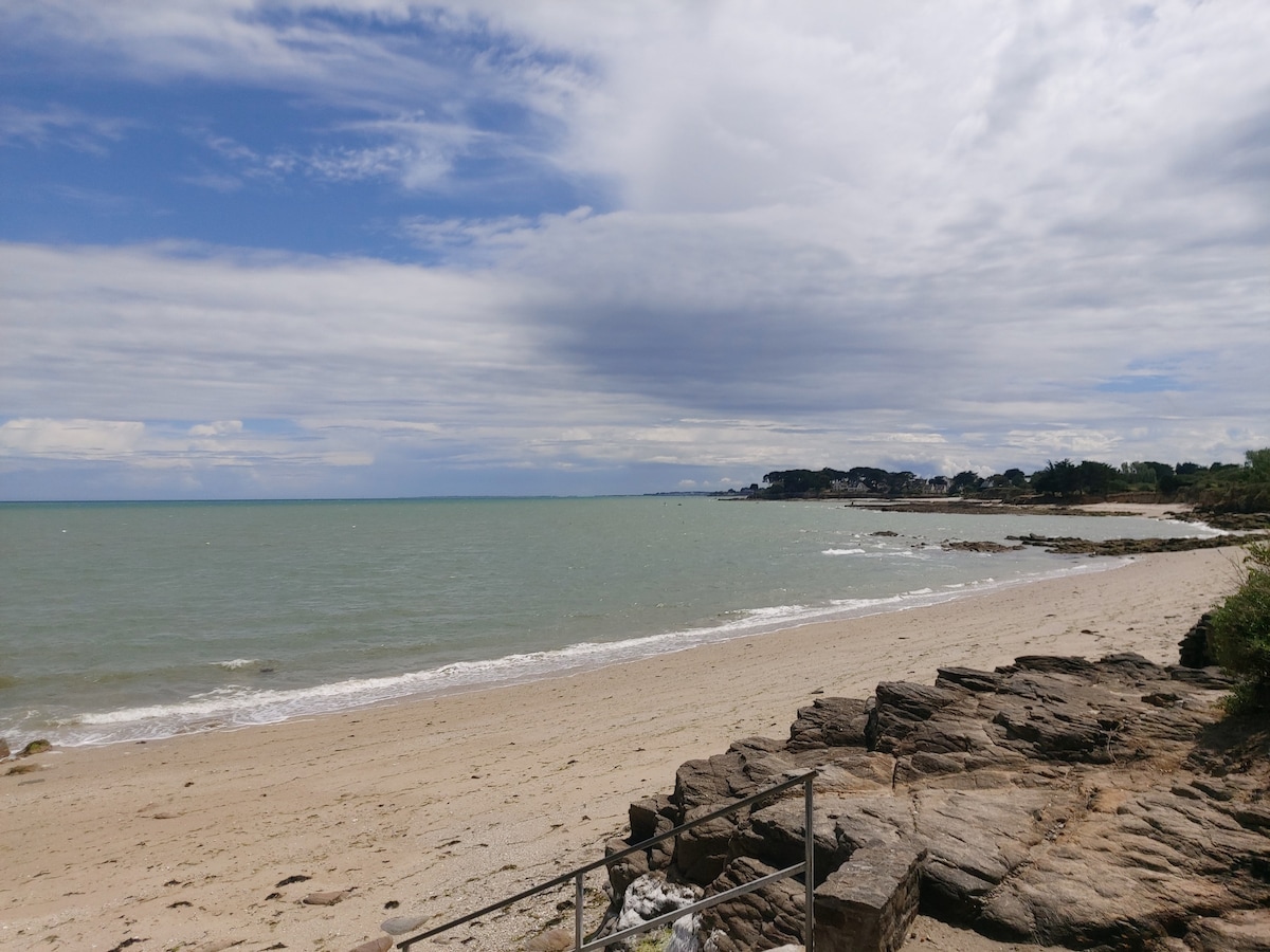 The image captures a serene beach scene with gentle waves lapping against the shore. A sandy stretch is visible, bordered by rocky formations. In the distance, greenery and coastal structures can be seen against a cloudy sky, creating a peaceful atmosphere.