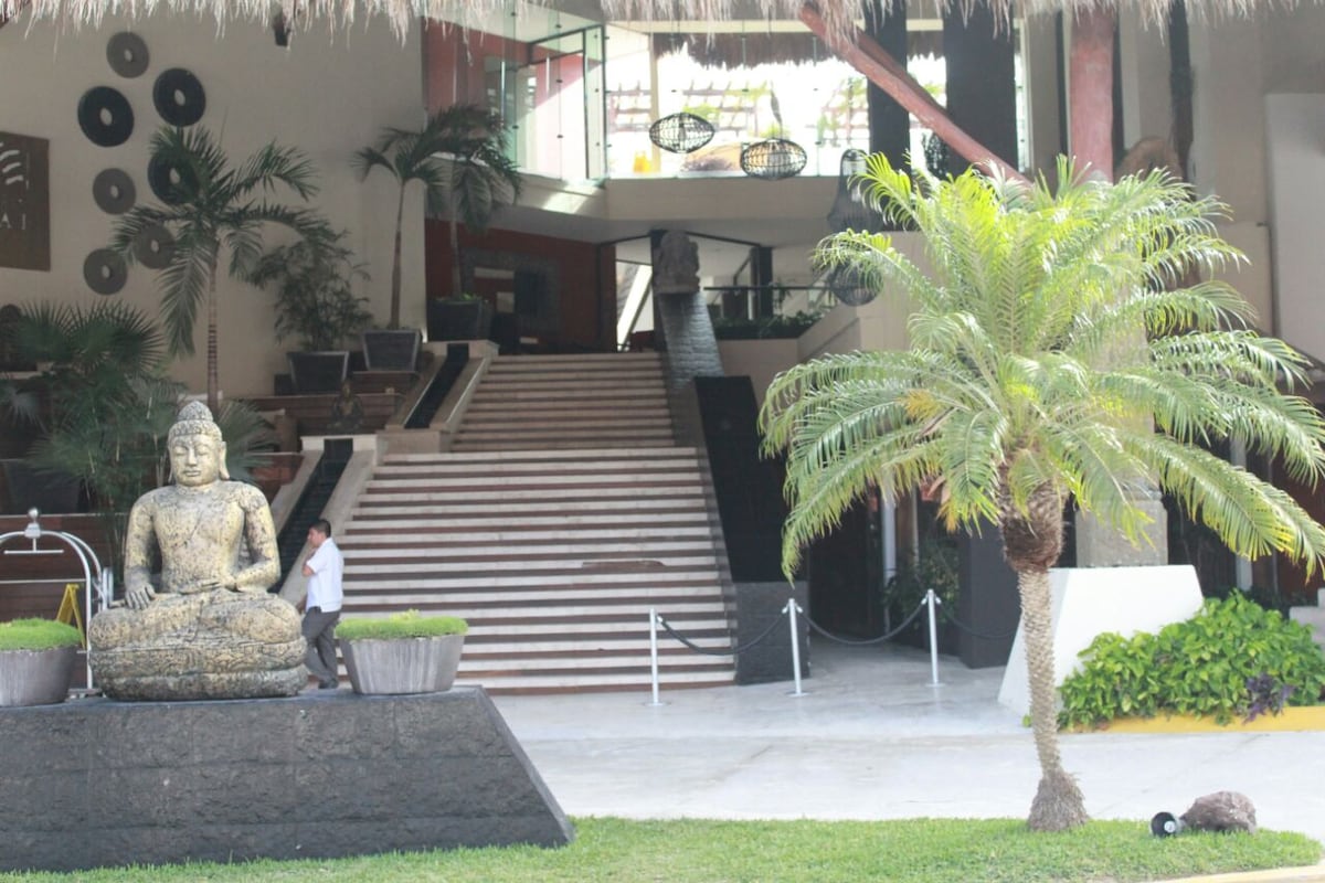 A grand entrance showcases a statue of Buddha, flanked by lush palm trees. A staircase leads to an upper level, surrounded by tropical plants, while modern decor elements enhance the serene environment. The open space invites guests into a tranquil atmosphere.