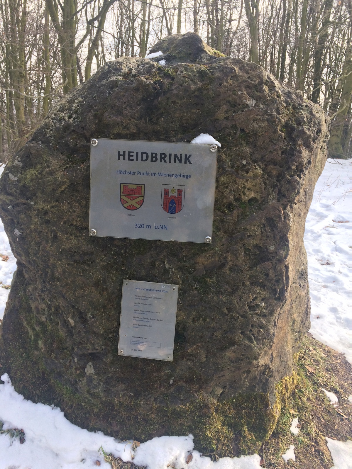 A large, moss-covered rock is displayed prominently, featuring a metal plaque with the name 'Heidbrink' and local insignia. Snow dusts the surrounding area, while trees provide a natural backdrop, signifying the highest point in the Wiehengebirge.