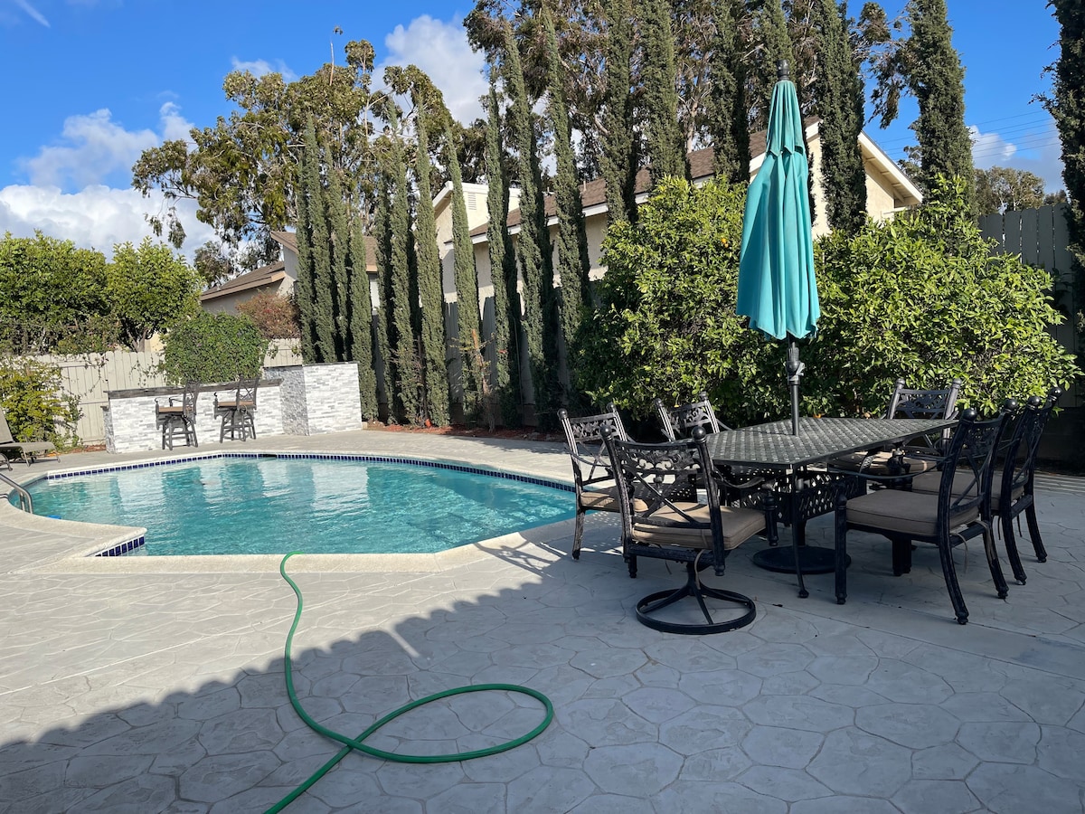 An outdoor space features a private swimming pool surrounded by a stamped concrete patio. A large outdoor dining table with multiple chairs is positioned nearby, complemented by a large umbrella providing shade. Lush greenery and tall trees create a natural privacy barrier.