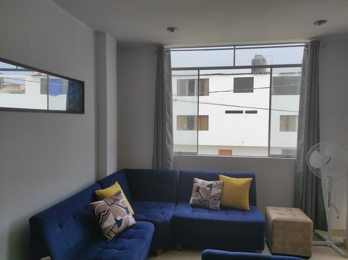 A bright living area is presented with a blue sectional sofa adorned with patterned cushions. Large windows allow natural light, with light gray curtains framing the view of the exterior. A small ottoman and a wall-mounted mirror complete the space.