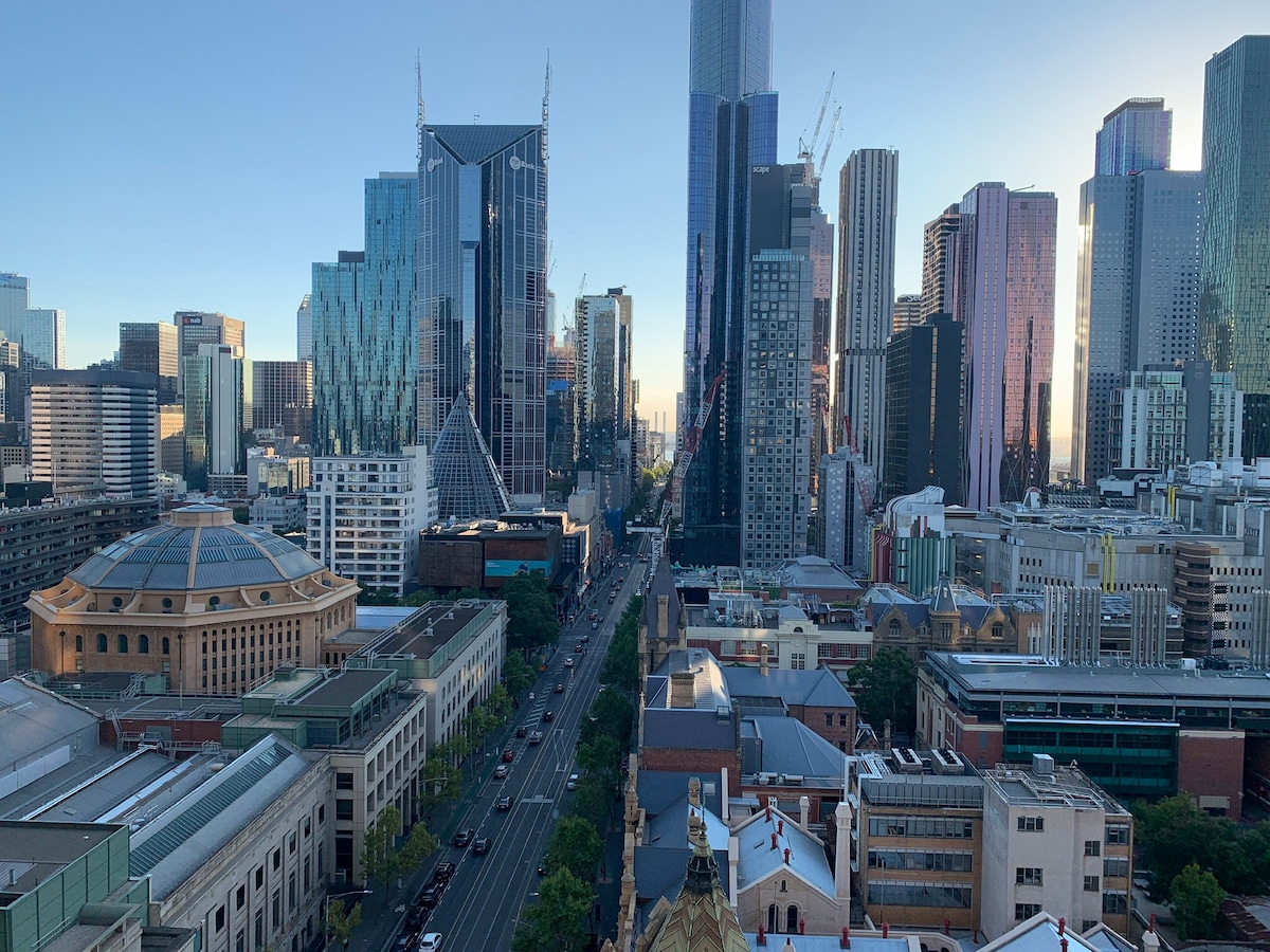 A panoramic view of Melbourne's skyline showcases a mix of modern skyscrapers and historical buildings. Streets lined with trees lead into the city, while construction cranes add a dynamic element to the vibrant urban setting under a clear blue sky.