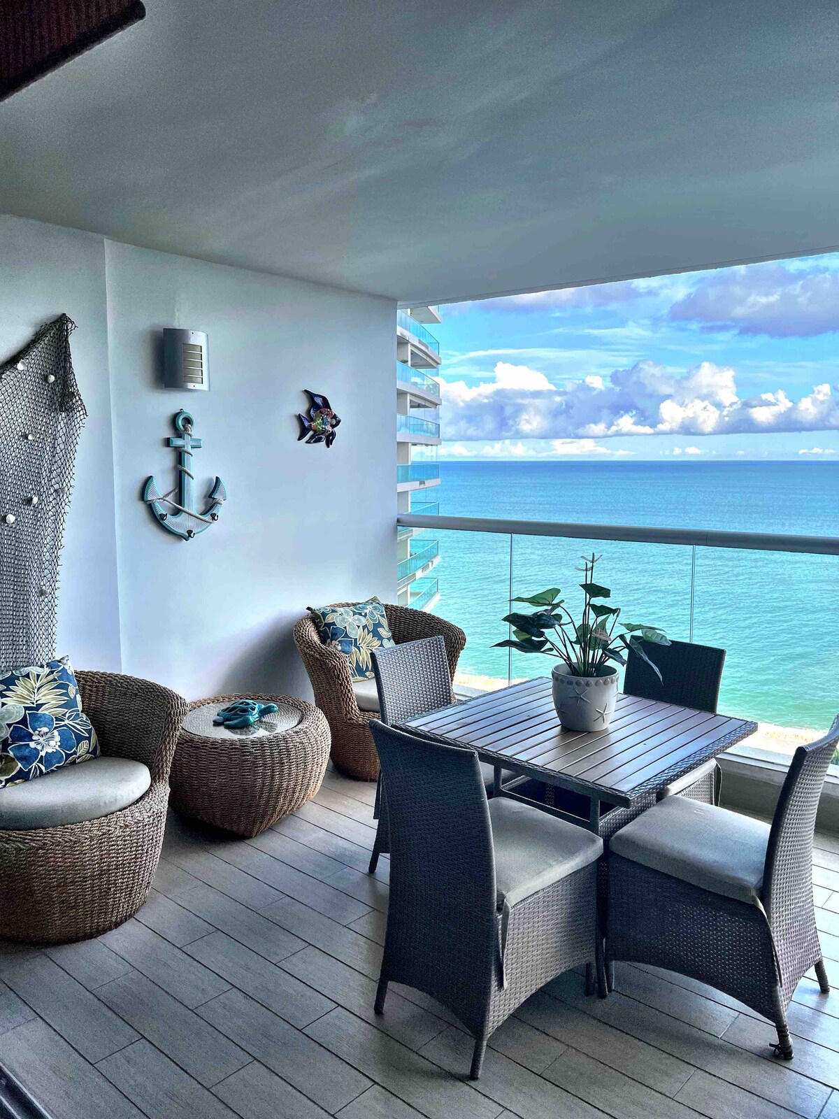A balcony area offers a table surrounded by four chairs, overlooking the ocean. Two cozy wicker chairs with cushions provide additional seating. Decorative elements, including an anchor and a wall hanging, add character to the space, which is illuminated by natural light.