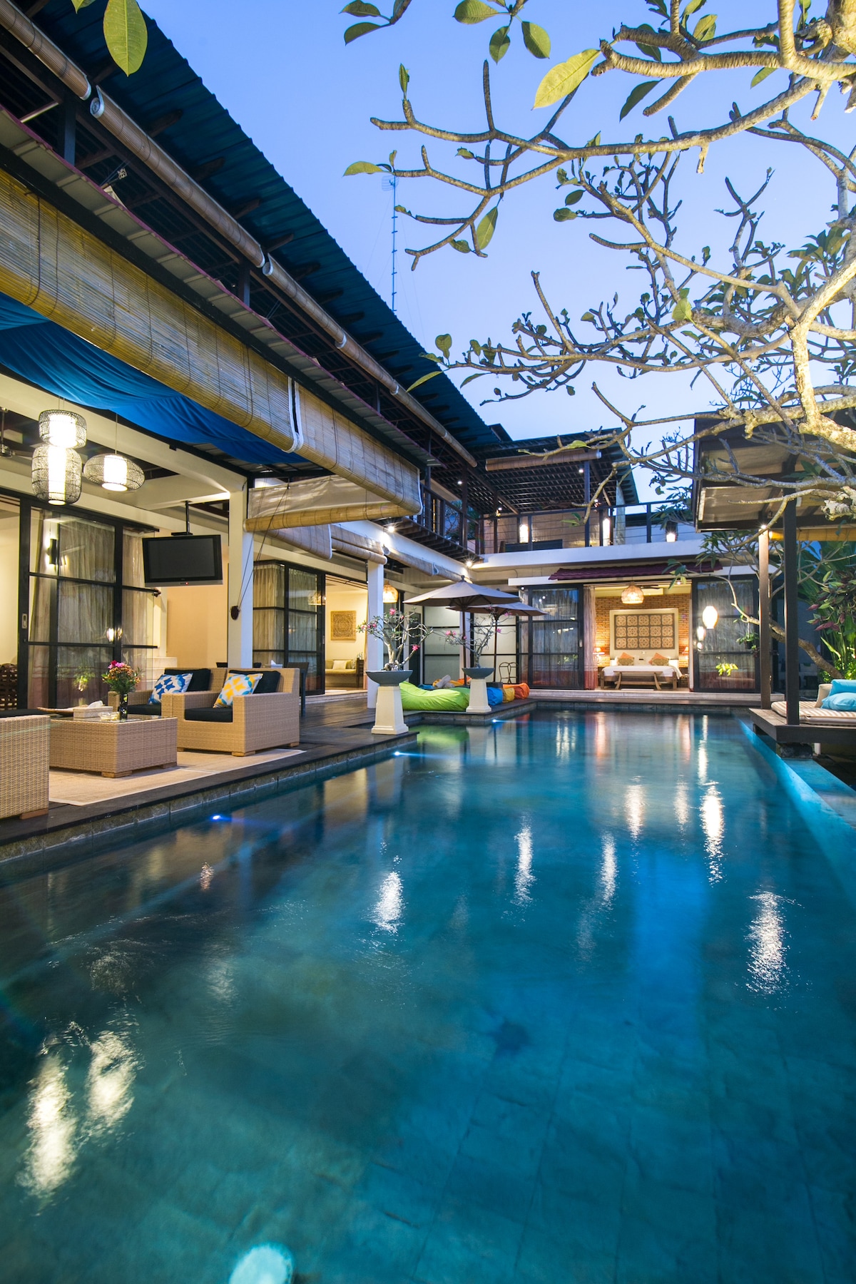 A tranquil outdoor space is featured, showcasing a clear swimming pool surrounded by modern Balinese architecture. Comfortable seating areas are positioned along the poolside, with vibrant cushions adding color. Lush greenery and elegant lighting create a welcoming ambiance as twilight approaches.
