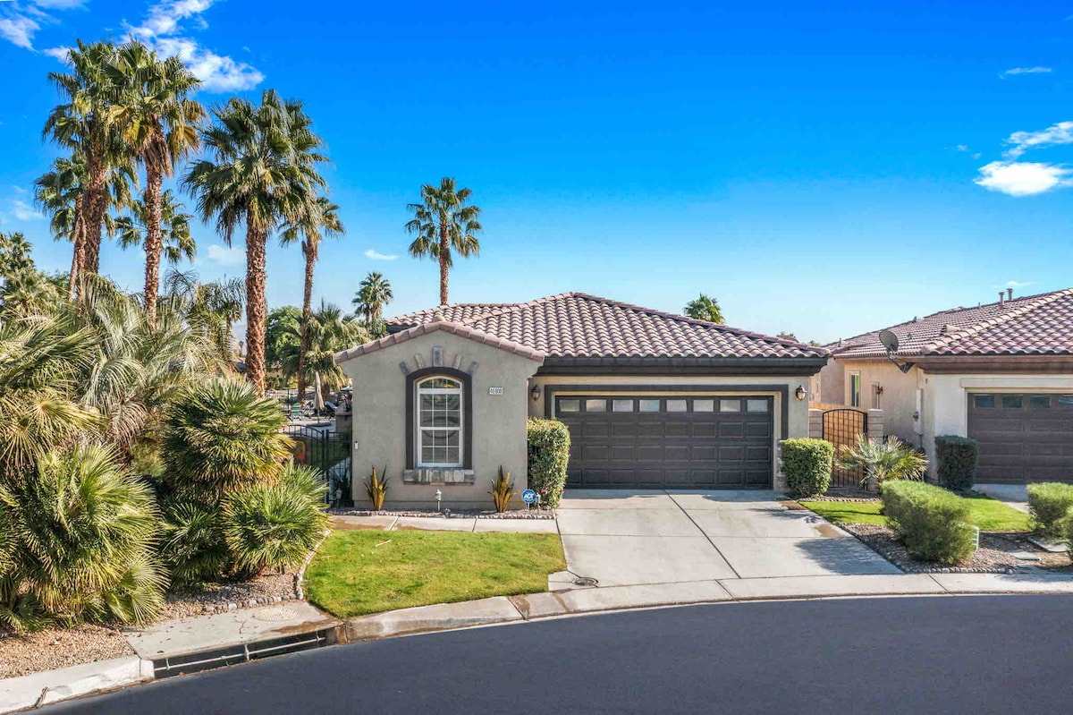 The front view of a single-story home is presented, featuring a tiled roof and a two-car garage. Lush palm trees and desert landscaping enhance the welcoming appearance. The driveway leads to a meticulously maintained lawn and adjacent homes are visible in a gated community setting.