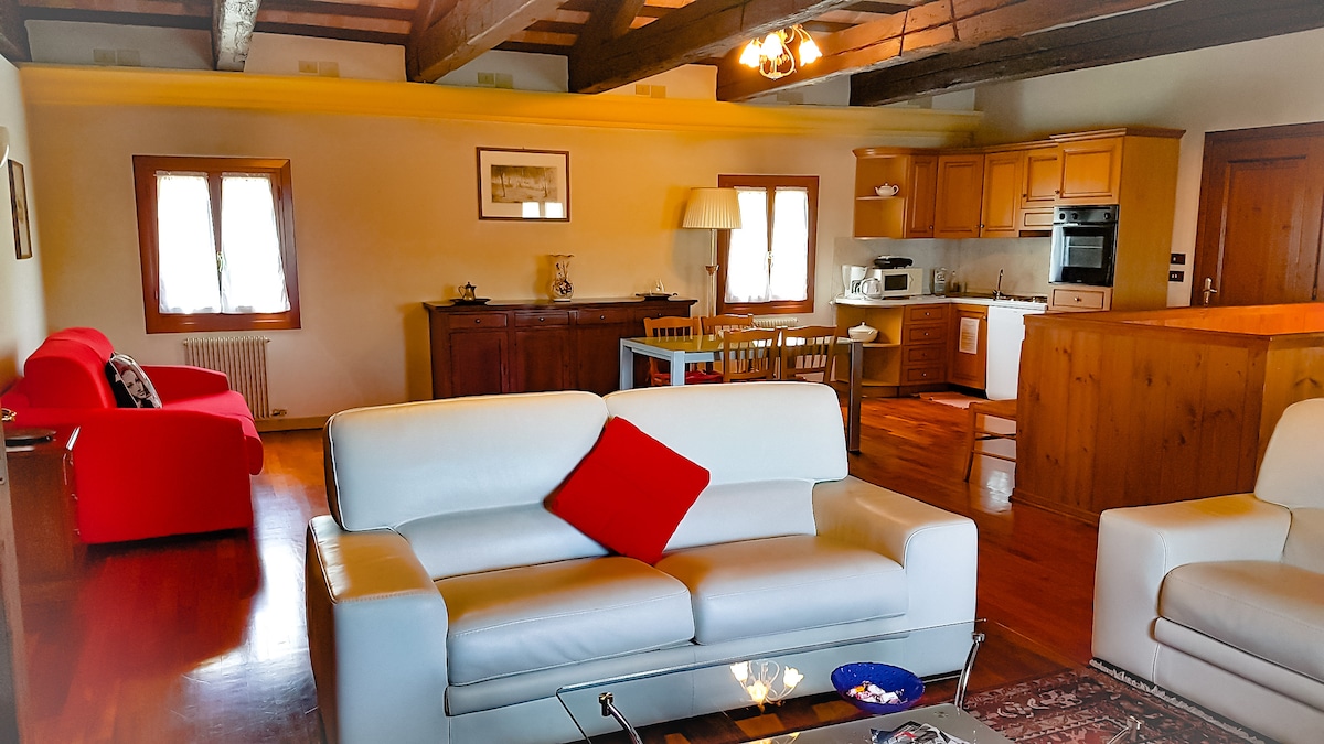 An open space living area is showcased, featuring a modern white sofa with a red accent pillow and a red chair. A wooden dining table is present near the kitchen area, which is equipped with cabinetry and appliances. Natural light enters through multiple windows, enhancing the warm wood flooring.