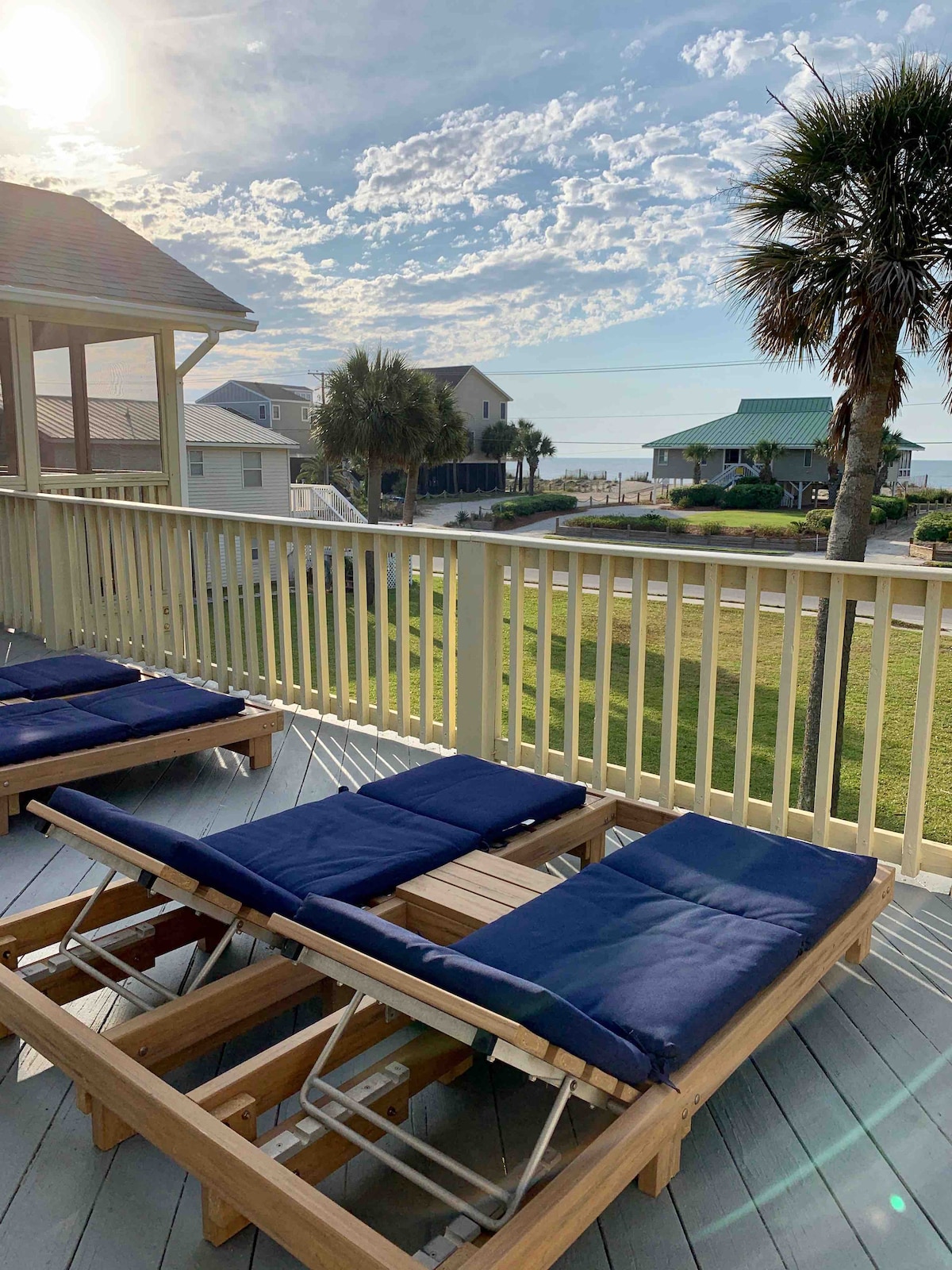 A spacious deck features multiple lounge chairs with blue cushions, providing a relaxing spot to enjoy ocean views. The horizon displays a vibrant sky, while nearby palm trees and beach houses contribute to the coastal atmosphere.