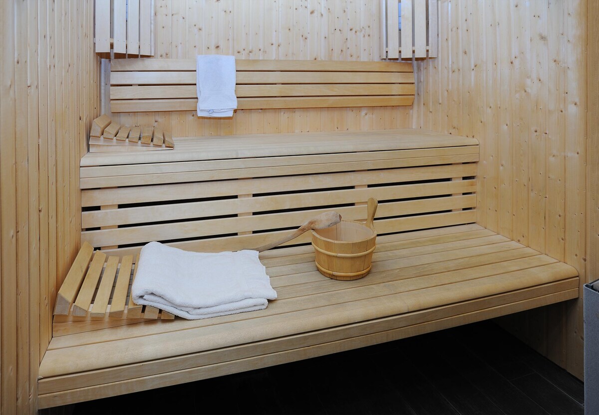 A wooden sauna space is shown, featuring two bench levels made of slatted wood. A light-colored towel and a wooden scoop are placed on the lower bench. The walls are lined with smooth wood panels, creating a warm and inviting environment.