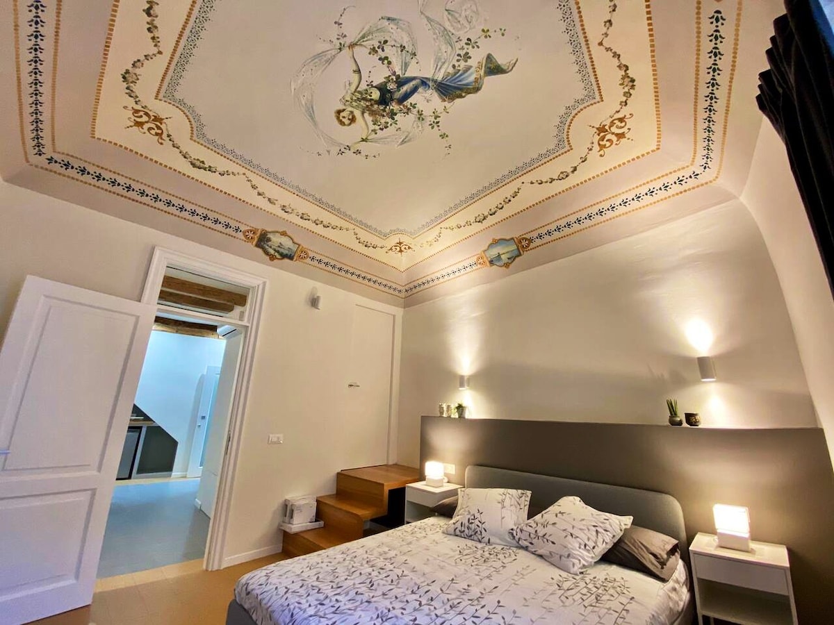The bedroom features a spacious bed with decorative pillows and bedside lamps. An intricately painted ceiling adds historical charm. A small set of elegant steps leads to another room, enhancing the space's unique layout.
