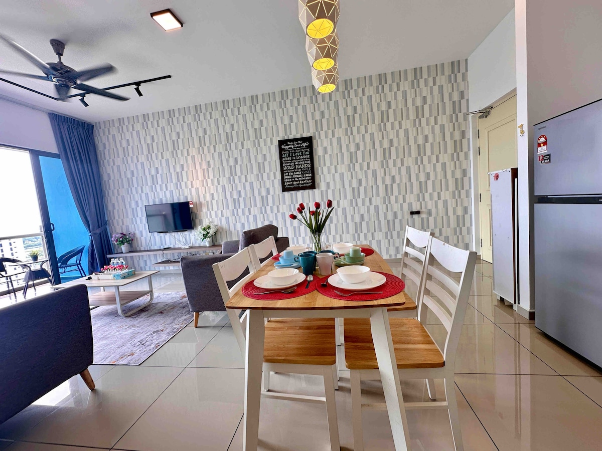 A dining area features a wooden table set for four, accompanied by white chairs. A modern kitchen with stainless steel appliances is visible in the background. Natural light floods the space through large windows, and a decorative wall adds an appealing texture.
