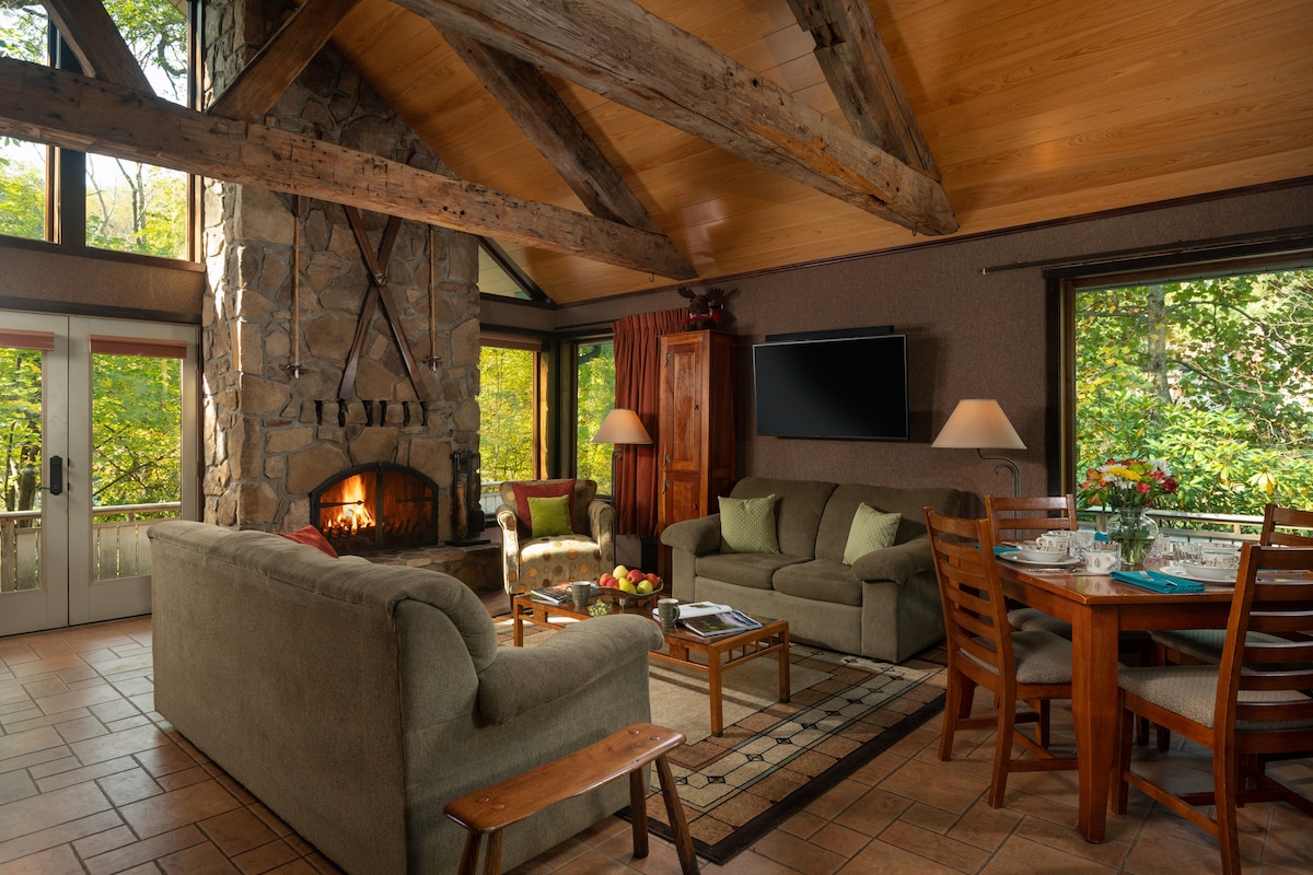 A spacious common room features a large fieldstone fireplace surrounded by comfortable seating. Large windows allow natural light to fill the space, showcasing views of the surrounding greenery. A wooden dining table is set for meals, inviting guests to gather and enjoy together.