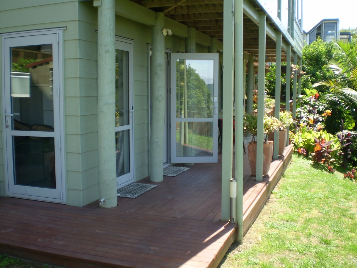 A private deck area is shown, featuring a combination of wooden flooring and green grass. Two glass door entrances provide access to the apartment, while planters filled with various plants enhance the garden setting.