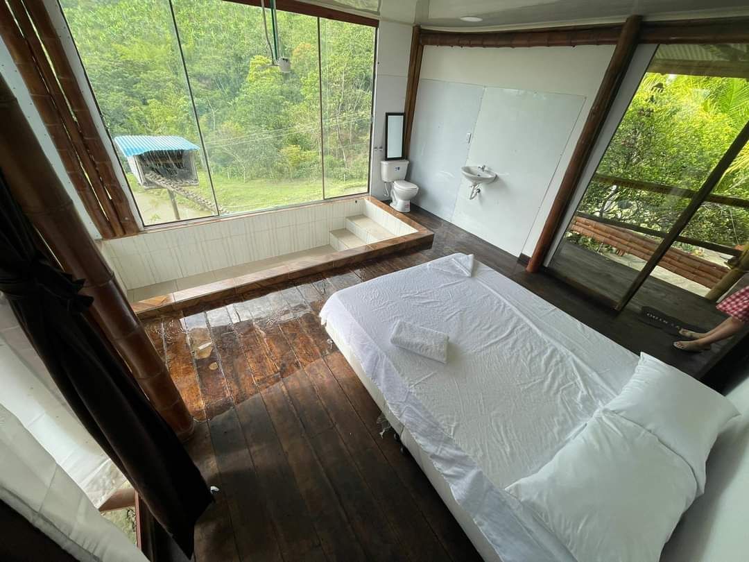 A room features large windows overlooking lush greenery, allowing natural light to fill the space. A spacious bed is presented with fresh linens. An en-suite bathroom with a tub and modern fixtures is visible, providing a tranquil and functional setting.