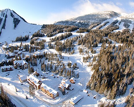 An aerial view captures a snow-covered landscape of the Red Resort area, featuring residential buildings nestled among trees. Ski slopes wind through the mountains in the background, indicating accessibility to outdoor activities. The bright white snow contrasts against the earthy tones of the buildings and forest.