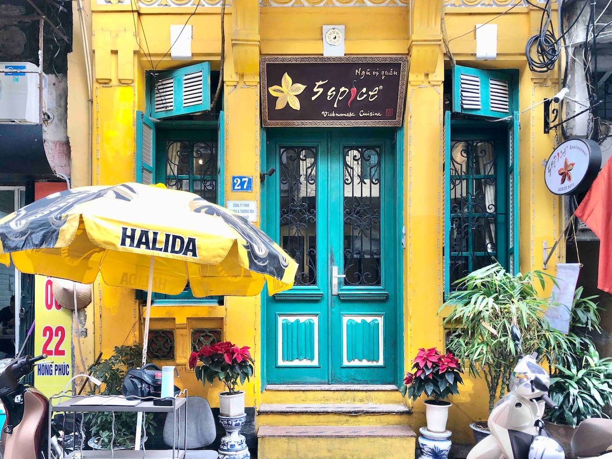 A charming building features bright yellow walls and teal doors framed by vibrant green plants. A small table with chairs is located in front, accompanied by a yellow umbrella displaying the word 'HALIDA.' The restaurant's sign enhances the inviting appearance of the entrance.