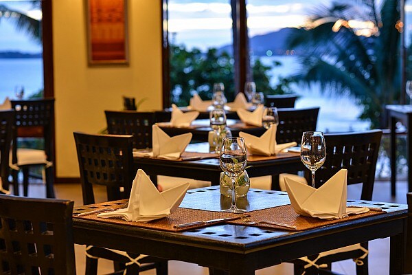 An elegant restaurant setting is showcased, featuring dark wooden tables arranged for dining with crisp white napkins neatly folded. Glass water bottles and wine glasses are placed at each setting. Large windows provide a view of the sea and palm trees beyond, enhancing the serene atmosphere.