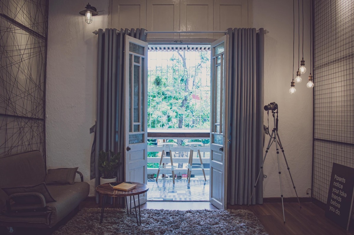 A set of double doors is opened to reveal a balcony with greenery visible outside. Simple curtains frame the doors, and a relaxed seating area with a small table is present. A decorative wall fixture and a tripod for photography are placed nearby.