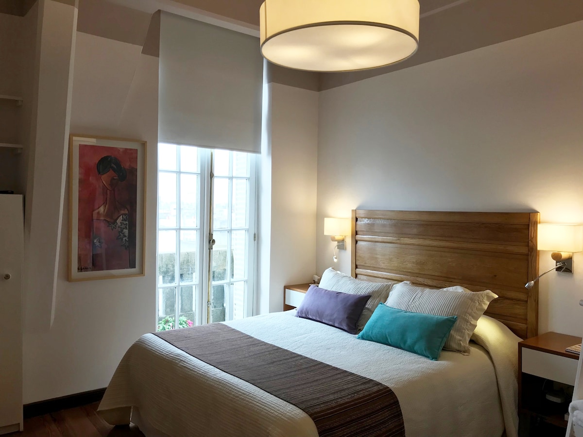 A well-appointed bedroom features a spacious bed adorned with a neutral-coloured blanket and decorative pillows in shades of turquoise and purple. Natural light enters through a large window, complemented by soft window coverings. A piece of wall art adds a touch of personality to the room.