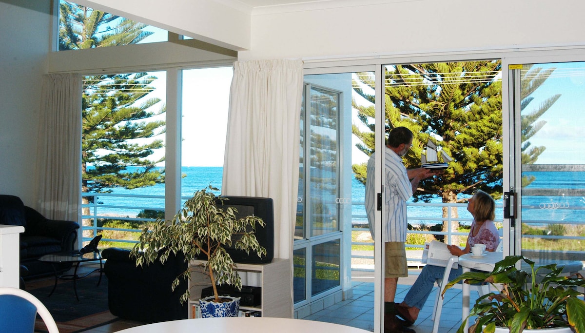 An open-plan living area offers large glass doors providing access to the seafront views. A dining table sits in the foreground, while a relaxed seating space is visible, complemented by indoor plants. The ocean and pine trees are framed beautifully outside.