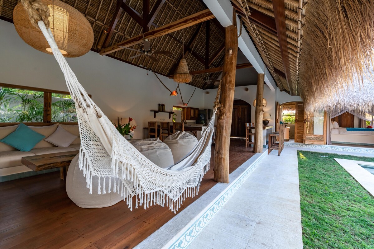 An open-air living space features a comfortable hammock suspended between wooden posts. Cozy seating areas with plush cushions are arranged nearby, and natural light filters through expansive windows, illuminating the adjoining kitchen and dining area. Lush greenery is visible in the background.