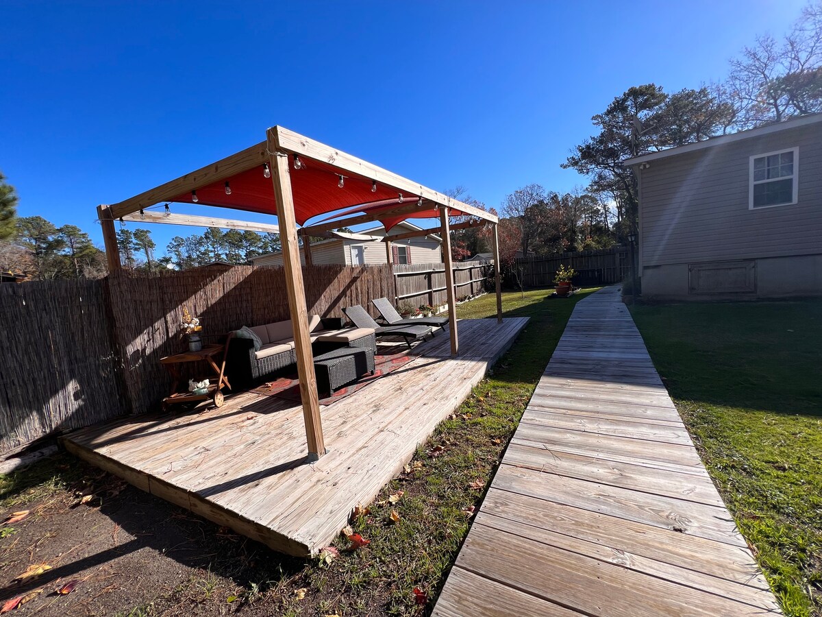 A wooden deck is shown, featuring a shaded seating area with comfortable furniture. A pathway leads from the deck to a green yard, with a wooden fence bordering the space. The sky is clear and blue, enhancing the outdoor ambiance.