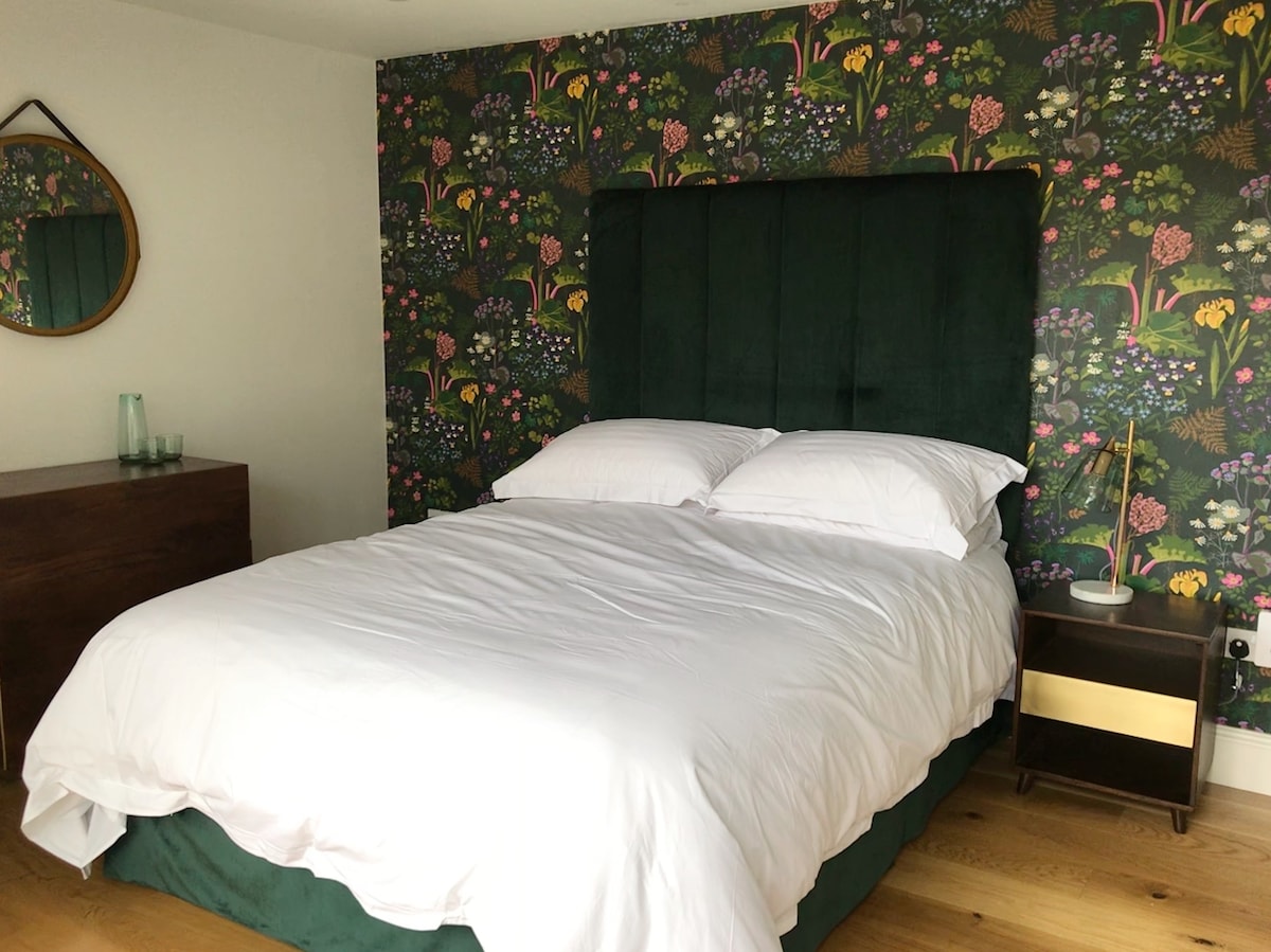 A cozy sleeping area features a spacious bed with crisp white linens, complemented by a dark green headboard. A vibrant floral wallpaper adds a touch of color, while a circular mirror hangs above a stylish wooden nightstand, creating a harmonious and soothing environment.