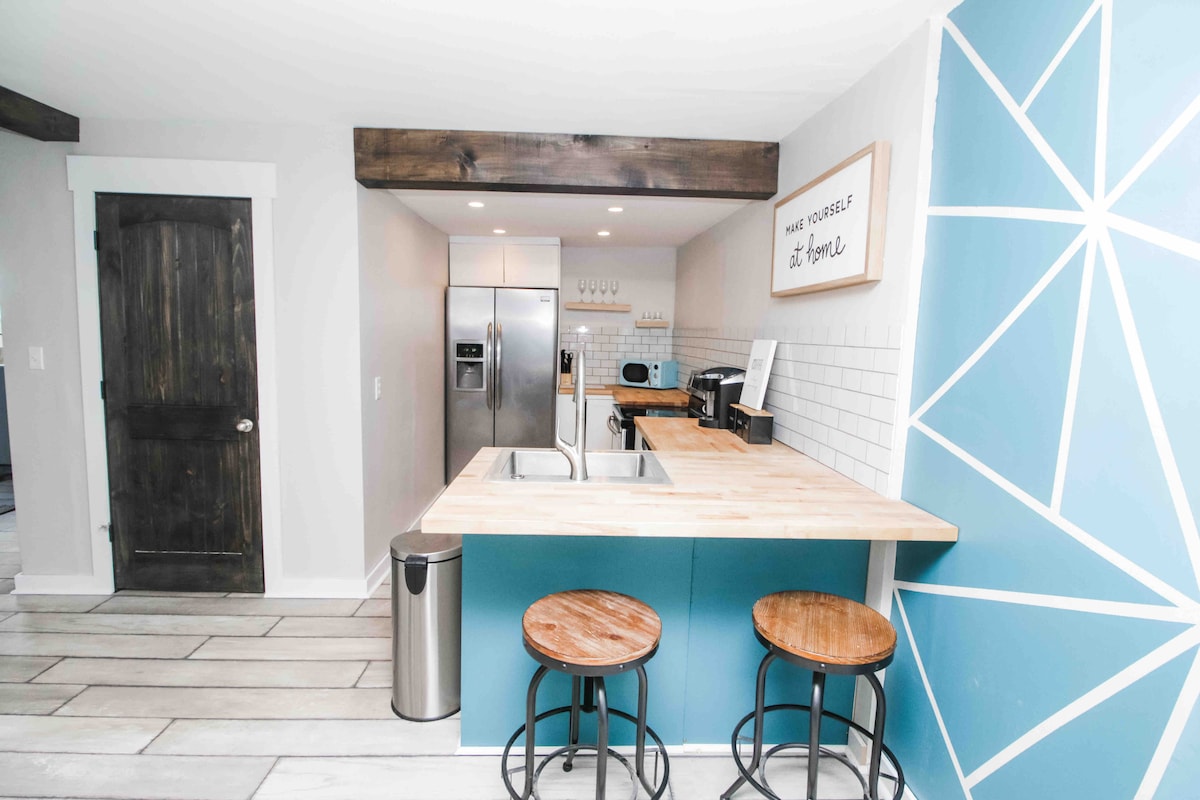 A modern kitchen space showcases butcher block countertops paired with stainless steel appliances. A vibrant blue accent wall features a geometric design. Two wooden stools are positioned at the kitchen island, complemented by white subway tiles that create a clean and inviting atmosphere.