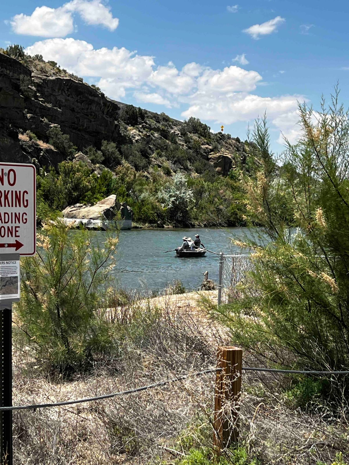 A serene river scene is depicted, featuring two individuals boating on calm water surrounded by lush greenery and rocky cliffs. A sign indicating no parking is visible in the foreground, and a natural landscape completes the tranquil setting.