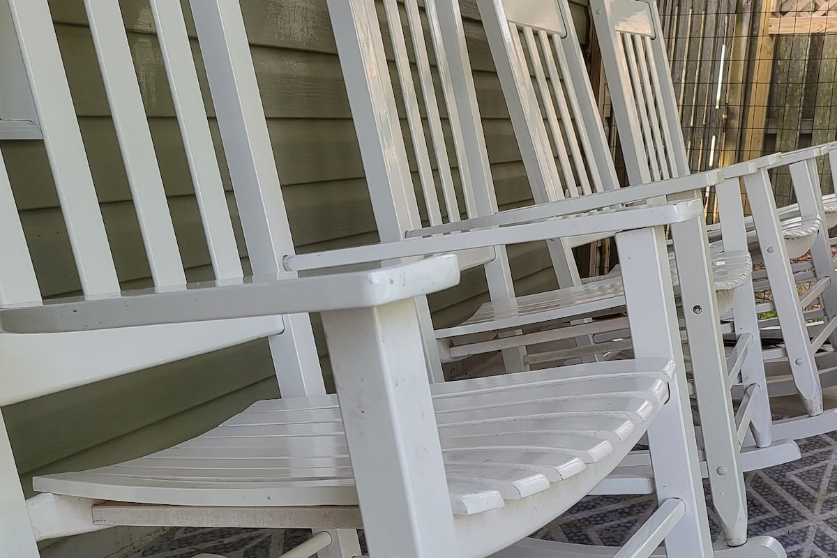 A row of white rocking chairs is positioned along a covered porch, providing an inviting seating area. The chairs feature classic slatted designs and are arranged to face a calm outdoor space, perfect for enjoying the surrounding atmosphere.