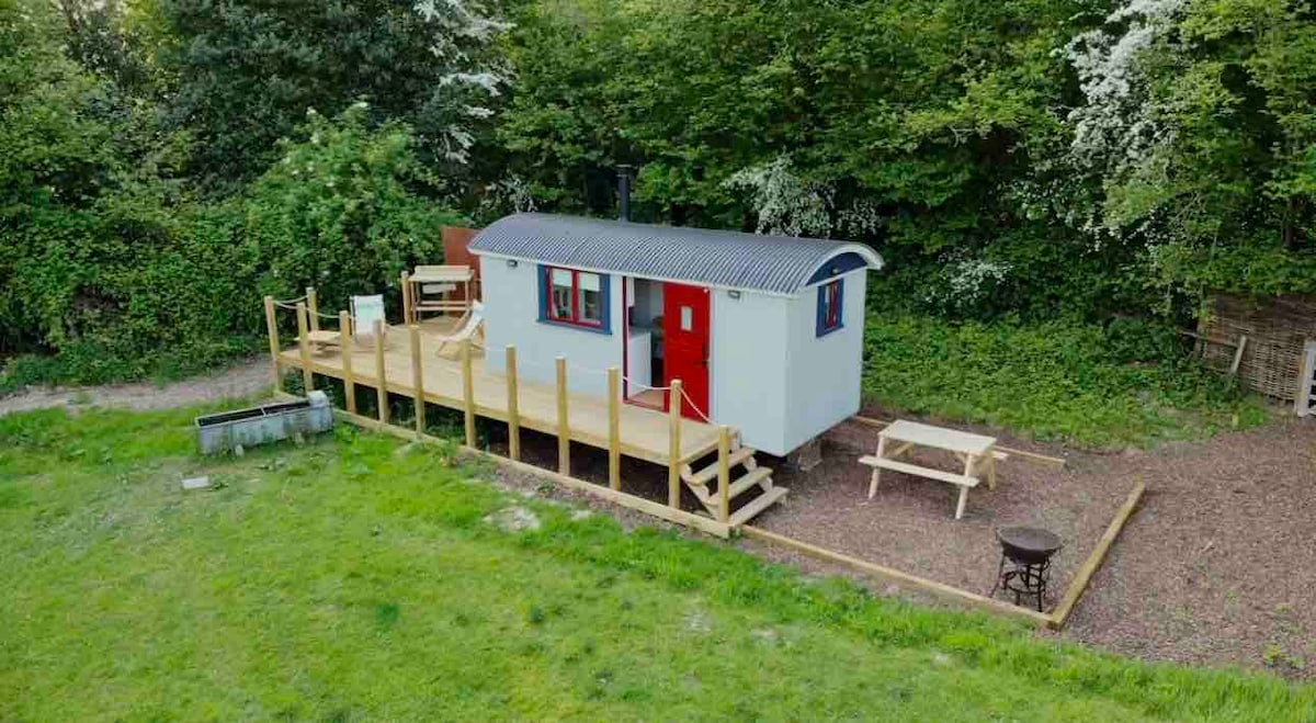 A shepherd's hut is set against a backdrop of greenery, featuring a wooden deck with several chairs and a picnic table. The structure displays a blend of red and grey exterior, complemented by large windows. Surrounding grass offers a serene, natural setting.