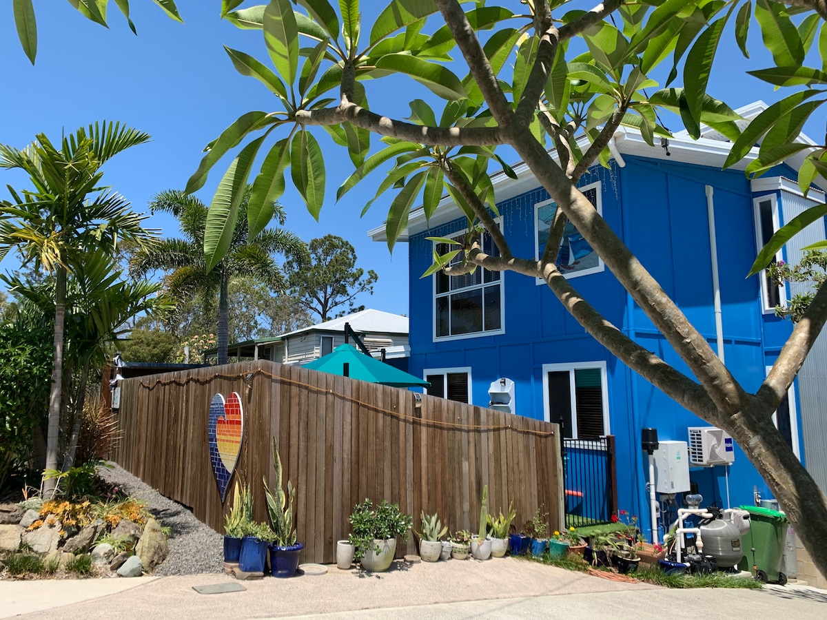 A vibrant blue exterior of the beach house is framed by tropical plants and a wooden fence. A rustic heart-shaped art piece is mounted on the fence, adding a colorful accent. Nearby, a private outdoor area is visible, inviting relaxation and enjoyment of the coastal surroundings.