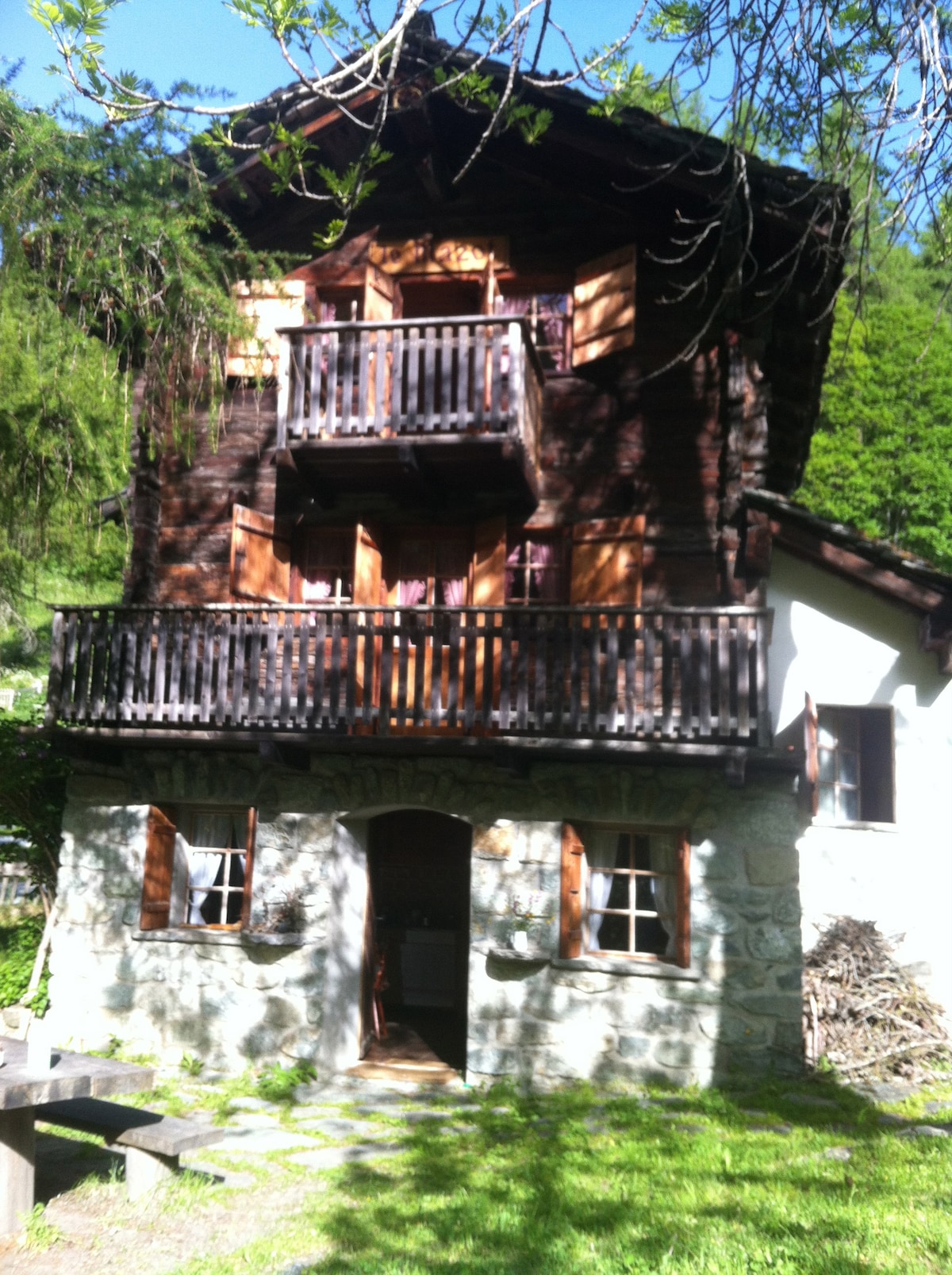 The chalet features a charming wooden facade, accentuated by balcony railings. Large windows allow natural light to enter, while a stone base adds rustic appeal. Surrounded by greenery, the entrance path is lined with grass, leading to the welcoming front door.