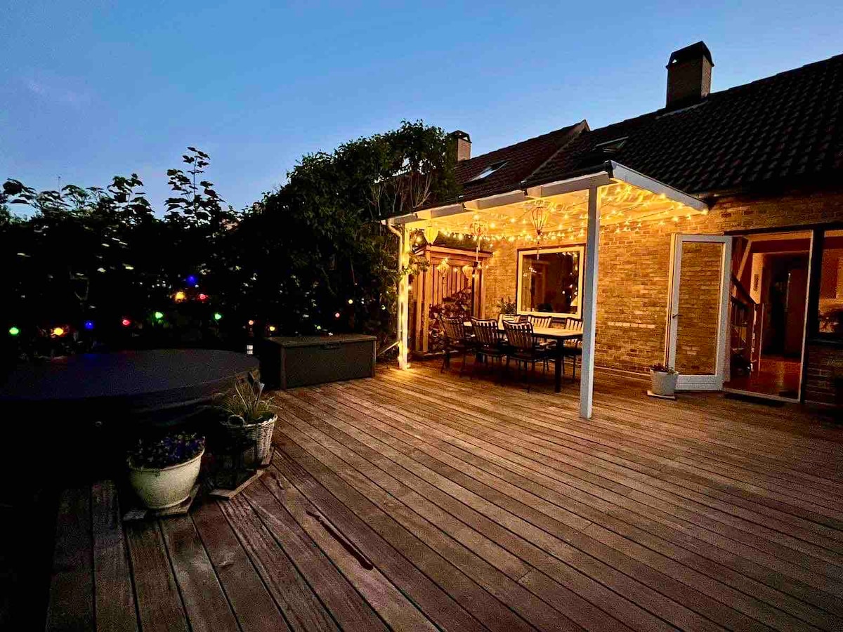A spacious wooden terrace is shown, illuminated by soft outdoor lighting. A large dining table with multiple chairs is positioned under an overhead structure. A cozy hot tub is visible alongside potted plants, surrounded by greenery that enhances the outdoor area.