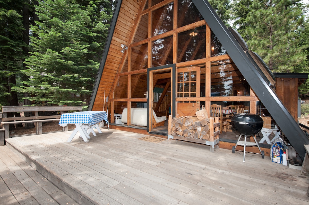 The cabin's exterior presents a striking A-frame structure with expansive glass windows. A wooden deck features a picnic table covered with a blue checkered cloth, while a charcoal grill is positioned nearby. Stacked firewood is neatly arranged in a designated area.
