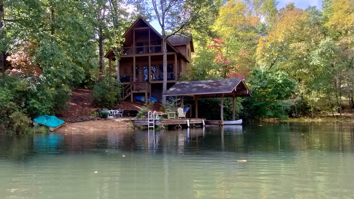 A waterfront home is nestled among trees, featuring wood accents and large windows. A private dock with seating extends into the calm lake, accompanied by canoes and kayaks. Soft green hues of nature surround the scene, creating a serene outdoor environment.