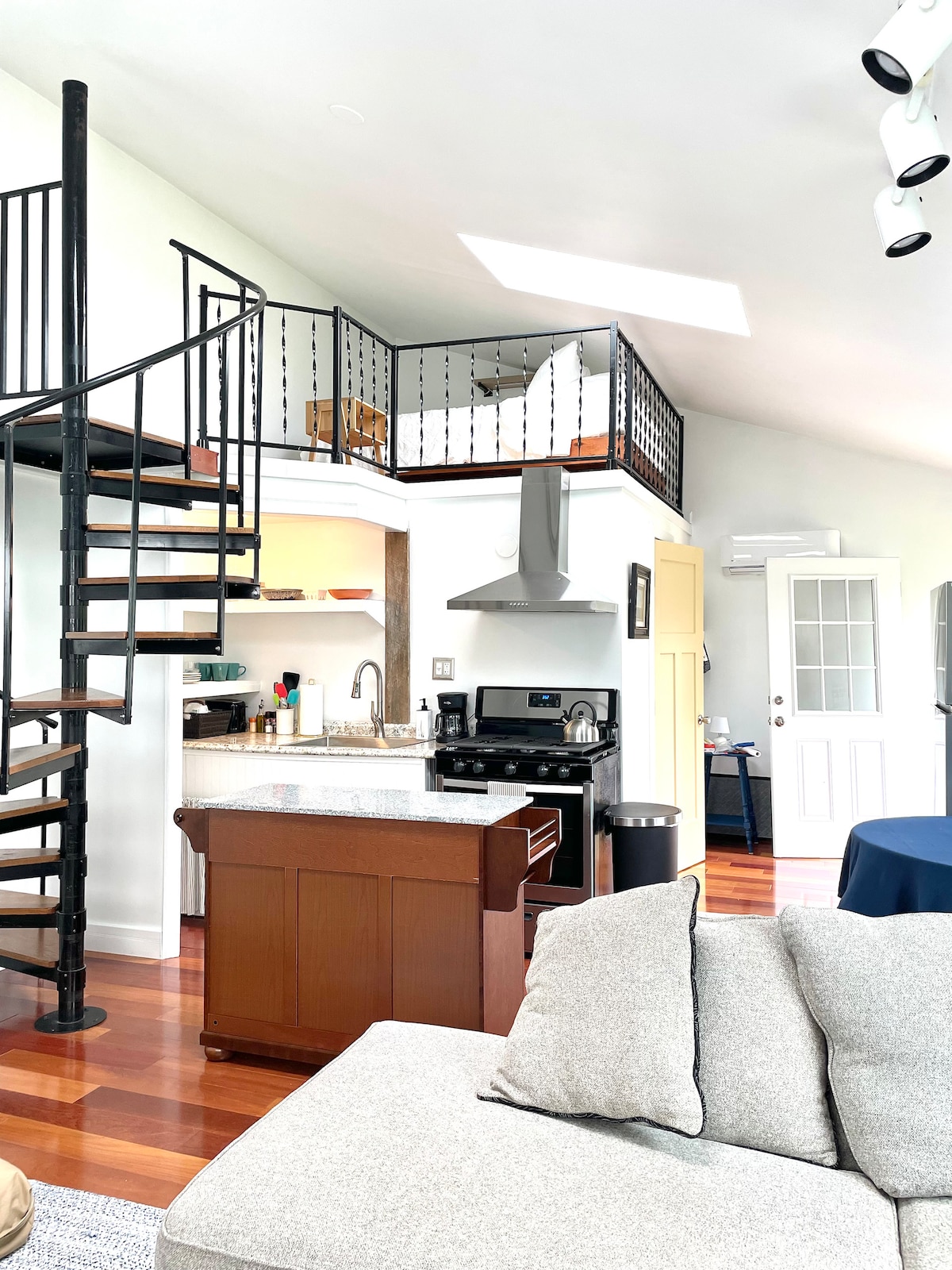 A spiral staircase leads to a small loft area, while the kitchen features a gas range and a full-size refrigerator. The open layout allows for light to fill the spacious living area, which includes a sectional couch and a dining area with a table.
